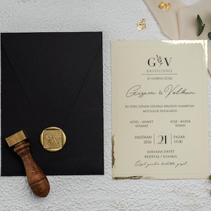 May include: A black envelope with a gold wax seal and a wooden stamp. A cream-colored invitation card with gold accents and text is next to the envelope. The invitation has the text "Gizem & Volkan".
