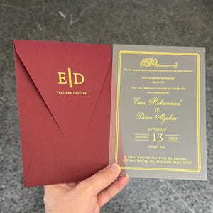 May include: A wedding invitation suite. The burgundy envelope has gold lettering, and the translucent invitation card features gold text. The card includes the names Enos Mohammad & Dana Alzabin, the date, time, and venue details.