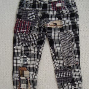 May include: Pants featuring a black and white plaid pattern with multiple visible patches in various checkered and striped fabrics. The garment includes frayed edges, exposed stitching, and white paint splatters across the surface.