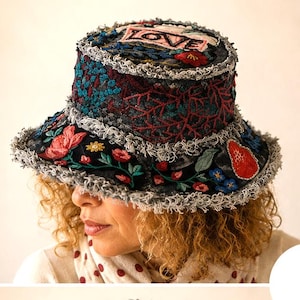Op de afbeelding: Een zwarte bucket hat met kleurrijk bloemenborduurwerk en het woord "LOVE" erop. De hoed heeft een gerafelde rand en is versierd met rode, blauwe en gele bloemen. Een uniek modeaccessoire.