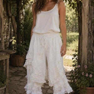 May include: A white sleeveless tank top paired with white cropped trousers featuring an elastic drawstring waistband, layered lace ruffles, and intricate floral embroidery details. The trousers have a wide-leg silhouette and are worn with brown boots.