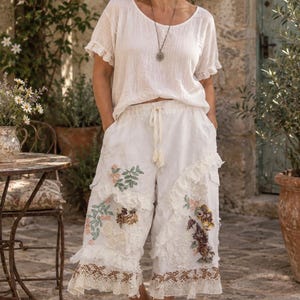 May include: A white short-sleeved top with ruffled edges and a round neckline, paired with white cropped trousers featuring a drawstring waist, layered lace ruffles, and floral embroidery patches in shades of green, pink, and brown.