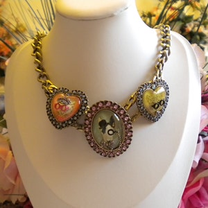 Può includere: Una collana a catena dorata con tre ciondoli a forma di cuore. I ciondoli presentano immagini colorate di animali e fiori.