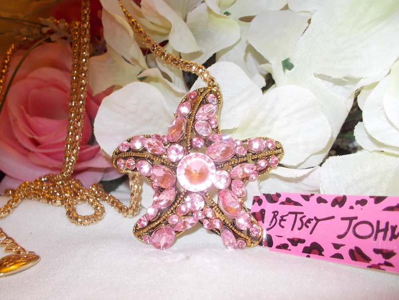 Peut inclure: Un collier dor&eacute; avec un pendentif en forme d'&eacute;toile de mer rose orn&eacute; de strass roses. Le pendentif a une pierre centrale et est pos&eacute; sur des fleurs blanches et roses. Une &eacute;tiquette rose avec "Betsey Johnson" est visible.