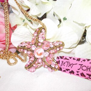 Peut inclure: Un collier dor&eacute; avec un grand pendentif en forme d'&eacute;toile de mer orn&eacute; de strass roses. Le pendentif est incrust&eacute; de strass roses et d'une pierre rose centrale plus grande. Le collier est pr&eacute;sent&eacute; avec des fleurs blanches et une &eacute;tiquette rose avec "Betsey Johnson."