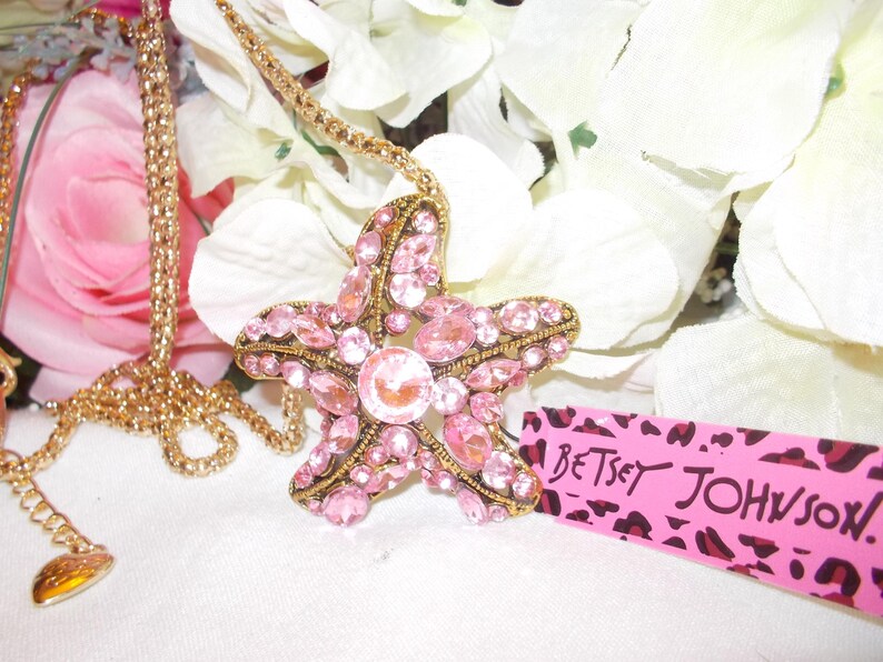 Peut inclure: Un collier dor&eacute; avec un grand pendentif en forme d'&eacute;toile de mer orn&eacute; de strass roses. Le pendentif est incrust&eacute; de strass roses et d'une pierre rose centrale plus grande. Le collier est pr&eacute;sent&eacute; avec des fleurs blanches et une &eacute;tiquette rose avec "Betsey Johnson."