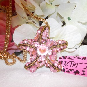 Peut inclure: Un collier dor&eacute; avec un pendentif en forme d'&eacute;toile de mer rose orn&eacute; de strass roses. Le pendentif a une pierre centrale et est pos&eacute; sur des fleurs blanches et roses. Une &eacute;tiquette rose avec "Betsey Johnson" est visible.