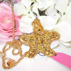 Peut inclure: Un collier dor&eacute; avec un pendentif en forme d'&eacute;toile de mer. Le pendentif pr&eacute;sente un design textur&eacute; et d&eacute;taill&eacute;, orn&eacute; de petits &eacute;l&eacute;ments scintillants. Le collier est pr&eacute;sent&eacute; sur un fond de fleurs roses et blanches.