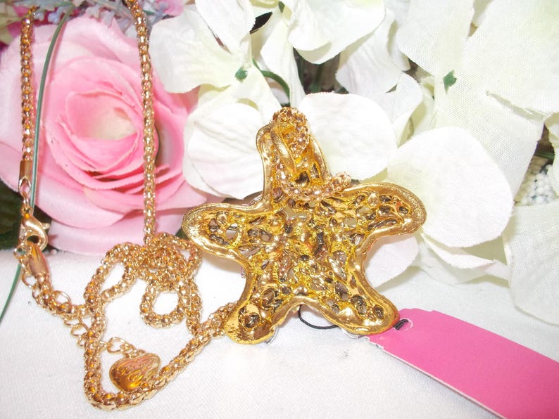 Peut inclure: Un collier dor&eacute; avec un pendentif en forme d'&eacute;toile de mer. Le pendentif pr&eacute;sente un design textur&eacute; et d&eacute;taill&eacute;, orn&eacute; de petits &eacute;l&eacute;ments scintillants. Le collier est pr&eacute;sent&eacute; sur un fond de fleurs roses et blanches.