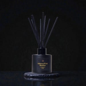 May include: A black diffuser bottle with black reeds, labelled "Tobacco & Oak Bourbon." The bottle sits on a dark, textured stone. The background is a solid black, creating a dramatic contrast.