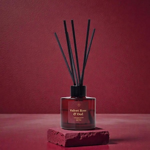 May include: A red glass diffuser bottle with black reeds, labelled "Velvet Rose & Oud." The bottle is on a red brick-like base against a textured red backdrop. The product is designed for home fragrance.