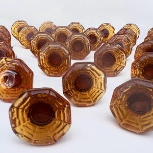 Pode incluir: Uma coleção de puxadores de vidro âmbar com formato octogonal. Os puxadores apresentam um padrão texturizado, semelhante a uma teia de aranha, e um centro marrom escuro. Os puxadores estão dispostos em fileiras, mostrando seu design e cor.