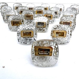 Peut inclure: Ensemble de boutons de placard carrés et transparents avec un aspect de verre martelé texturé. Chaque bouton présente un insert rectangulaire en métal doré. Les boutons sont disposés sur une surface blanche.
