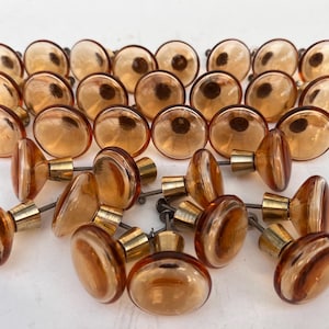 May include: A collection of amber-colored glass cabinet knobs with brass hardware. The knobs have an oval shape with a striped design. The knobs are arranged in rows and are suitable for furniture restoration or DIY projects.
