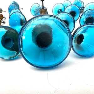 May include: A group of blue glass knobs with a shiny finish. The knobs are round and have a slight indentation in the center. They are arranged in a random pattern on a white surface.