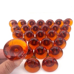 Könnte beinhalten: Eine Sammlung bernsteinfarbener Glasgriffe, angeordnet in einem dreieckigen Muster. Jeder Knopf hat eine abgerundete, kuppelförmige Form mit dunkler Mitte und einem kleinen Metallschaft. Die Knöpfe sind vor weißem Hintergrund ausgestellt.