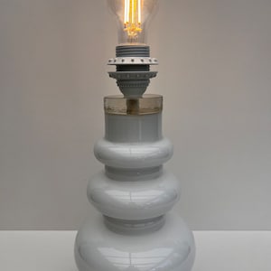 Könnte beinhalten: Eine weiße, moderne Tischlampe mit einem gestapelten, abgerundeten Sockel und einer durchsichtigen Glühbirne. Die Glühbirne leuchtet und strahlt ein warmes Licht aus. Die Lampe hat ein schlichtes, elegantes Design, geeignet für ein Schlaf- oder Wohnzimmer. Die Lampe ist etwa 38 cm hoch.