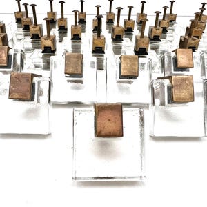 Op de afbeelding: Een opstelling van heldere glazen blokken met messingkleurige metalen accenten. Sommige blokken hebben een vierkant metalen stuk, andere een metalen stuk met een lange, dunne staaf die omhoog steekt. Gerangschikt op een witte ondergrond.