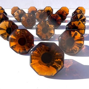 Può includere: Una collezione di 18 pomelli per armadi in vetro ambrato con forma ottagonale e design sfaccettato. I pomelli hanno un centro marrone scuro e sono disposti a forma di triangolo.