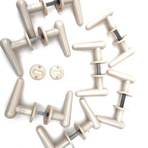 May include: A selection of off-white door handles, featuring a minimalist design. Each handle has a curved lever, a circular base, and a metal rod. Two keyhole covers are also visible. The handles are for doors.