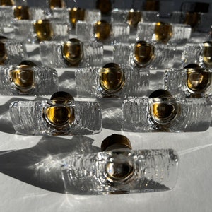 Pode incluir: Uma coleção de puxadores de armário de vidro transparente com uma aparência texturizada semelhante ao gelo. Cada puxador apresenta um detalhe metálico dourado e um topo escuro e arredondado. Os puxadores estão dispostos em fileiras, projetando sombras.