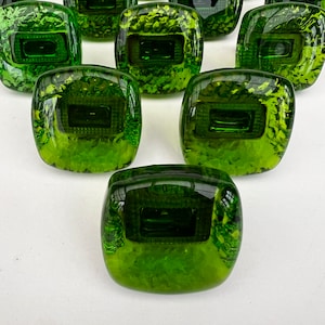 May include: A group of square, green glass objects with a textured, mottled appearance. Each piece has a rectangular indentation in the centre. The objects are arranged on a white surface, with some in focus and others blurred in the background.