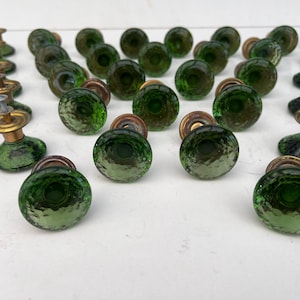 Op de afbeelding: Een verzameling vintage groene glazen deurknoppen met een getextureerd oppervlak en messing hardware. De knoppen zijn in rijen gerangschikt en tonen hun unieke ontwerp en kleur. Ideaal om een vleugje vintage charme toe te voegen aan elk decoratieproject.