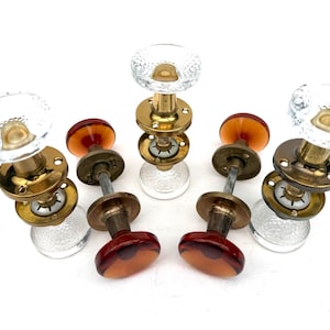 May include: A collection of five door knobs featuring three clear textured glass knobs with brass hardware and two amber-colored glass knobs with brass hardware. Each knob assembly includes a circular mounting plate and a central metal spindle.