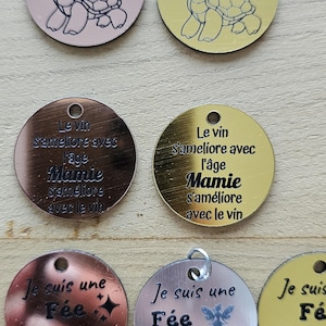 Peut inclure: Une collection de breloques rondes en métal avec divers motifs et textes. Certaines breloques présentent une illustration de tortue, tandis que d'autres comportent du texte français tel que "Le vin s'ameliore avec l'age Mamie". Les breloques sont dans des tons or, cuivre et argent.