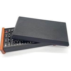 May include: A black synthesizer module with a wooden side panel and a black cover. The module features numerous knobs and switches. The cover is partially open, revealing the controls. The wooden side panel adds a touch of warmth to the design.