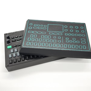 以下が含まれることがあります： 2つの黒い電子音楽制作デバイス。1つは、ターコイズ色の輪郭とボタンのグリッドを備えた長方形のパッドです。もう1つは、ノブ、ボタン、緑色のボタンを備えたコントロールサーフェスです。どちらのデバイスも白い表面にあります。