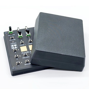 Può includere: Dispositivo audio elettronico nero con manopole e pulsanti, etichettato "VOLUME", "CHAOS", "TEMPO", "VOICE 1", "VOICE 2", "RUMBLE", "RECORD" e "ERASE". Un coperchio grigio scuro è parzialmente sopra il dispositivo. Lo sfondo è bianco.