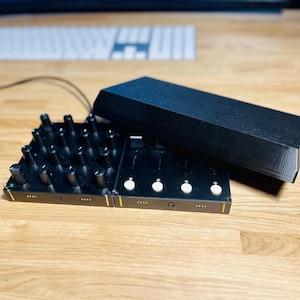 Könnte beinhalten: Ein schwarzer elektronischer Musikcontroller mit einem Raster aus Knöpfen und vier weißen Drehknöpfen. Der Controller liegt auf einer Holzoberfläche mit einem schwarzen Gehäuse darauf.