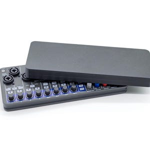 Puede incluir: Una mesa de mezclas de audio gris oscuro con una cubierta a juego. La mesa de mezclas tiene múltiples perillas, botones y conectores de entrada. Los botones están iluminados con luces azules. La cubierta está ligeramente entreabierta.