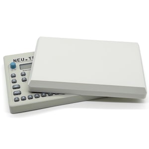 Puede incluir: Un dispositivo electrónico gris claro con teclado y pantalla, parcialmente cubierto por una cubierta rectangular blanca. El dispositivo tiene el texto "NEU. TI" impreso. La cubierta tiene un borde gris.