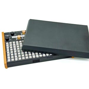 May include: Black electronic music production device with a grid of white buttons, black knobs, and a wooden side panel. A black cover is partially open, revealing the controls. The device is on a white background.