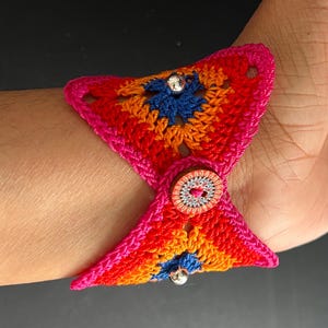 May include: A colorful crocheted bracelet with a button closure and silver beads. The bracelet is made with pink, orange, red, and blue yarn.