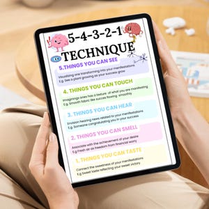 May include: A tablet displays a colorful infographic titled "5-4-3-2-1 TECHNIQUE." The graphic lists five sensory-based steps for manifestation, including things to see, touch, hear, smell, and taste. The tablet is held by a person.