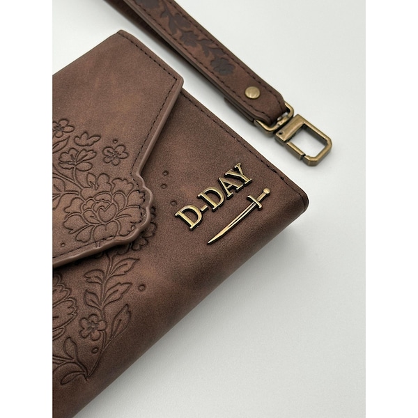 May include: A brown leather wallet with floral embossing and a matching wrist strap. The wallet features the text "D-DAY" in gold lettering with a sword emblem. The wrist strap has a floral pattern and a gold-colored clasp.
