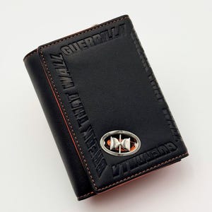 May include: Black leather wallet with the embossed text "GUERRILLA" and "BREAK THAT WALL". The wallet features orange stitching and a silver emblem with an orange center. The wallet is closed and has a tri-fold design.