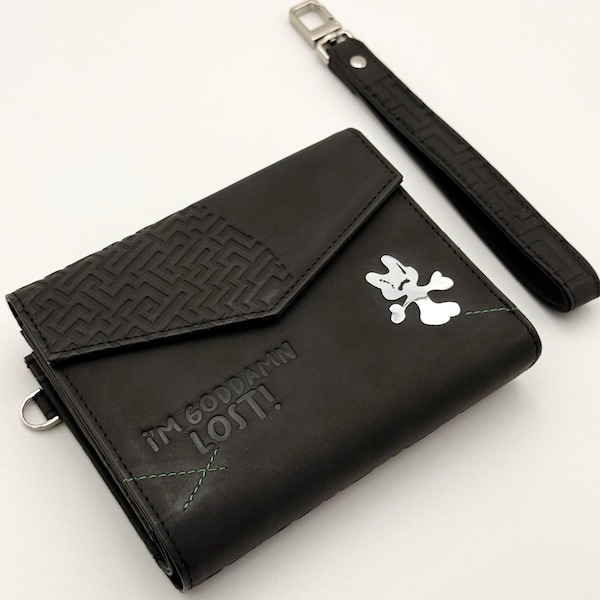 May include: A black leather wallet featuring a textured geometric flap, a silver metallic graphic of a character, and the embossed text I'M GODDAMN LOST! with green stitching details. Includes a matching detachable black wrist strap.
