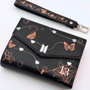 Puede incluir: Cartera negra con una correa de muñeca a juego. La cartera presenta un diseño floral y de mariposas en color cobre, junto con detalles de corazones blancos. El número "13" es visible en la parte delantera.