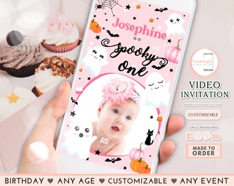 May include: A pink and black digital video invitation for a spooky one-year-old birthday party. The invitation features a photo frame with a baby girl wearing a pink headband and a ghost with the text "Boo!" on it. The text "Josephine is a spooky one" is written in black cursive font. The invitation also features a black cat, pumpkins, bats, and stars.