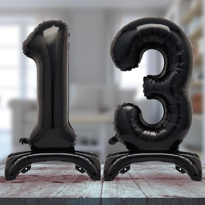 May include: Two black balloon numbers, 1 and 3, stand upright on a wooden surface. The balloons are inflated and have a shiny finish.