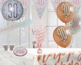 May include: A 90th birthday party decoration set with a rose gold theme. The set includes a banner, balloons, confetti, a table cover, and party plates and cups. The banner says "Happy 90th Birthday".