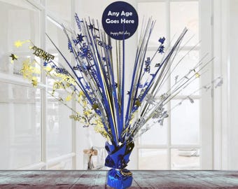 May include: Blue and silver birthday centerpiece with a blue circle that says "Any Age Goes Here" and "Happy Birthday".