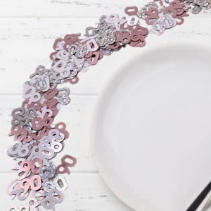 May include: A pile of silver and rose gold confetti shaped like the number nine. The confetti is scattered on a white wooden surface next to a white plate.