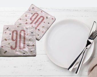 May include: Two white paper napkins with a pink "90" printed on them. The napkins have a confetti pattern with gold and brown dots. The napkins are on a white wooden surface.
