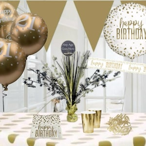 May include: Gold and white 21st birthday party decorations. The decorations include balloons, a banner, a table cover, and cups. The balloons are gold with white polka dots and the number 21 printed on them. The banner is white with gold letters that say "happy birthday". The table cover is white with gold polka dots. The cups are gold with a metallic finish.