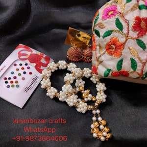 May include: A collection of jewellery and accessories displayed on a black satin background. The image features a white beaded necklace with gold details, red and gold earrings, a floral embroidered pouch, and a sheet of colourful bindi stickers.